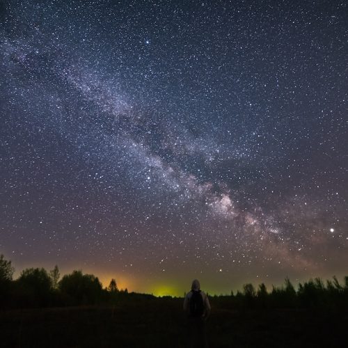 A man with a backpack standing at night, with his back to the camera, against the background of the Milky Way